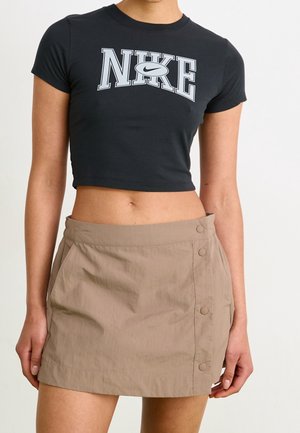 Osoba nosząca czarny krótki t-shirt Nike i beżową mini spódnicę z guzikami, stojąca z rękami swobodnie opuszczonymi wzdłuż ciała na jednolitym tle.