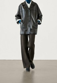 Person wearing black leather buttoned jacket, white shirt, blue turtleneck, dark wide-leg pants, and black textured shoes walking indoors.