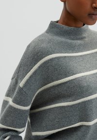 Pull en tricot gris avec un col montant, présentant des rayures horizontales crème, une texture douce et une coupe décontractée.