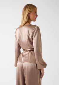 Top in raso a maniche lunghe color marrone chiaro con vita aderente e maniche svasate, caratterizzato da una texture morbida e un orlo posteriore curvo.