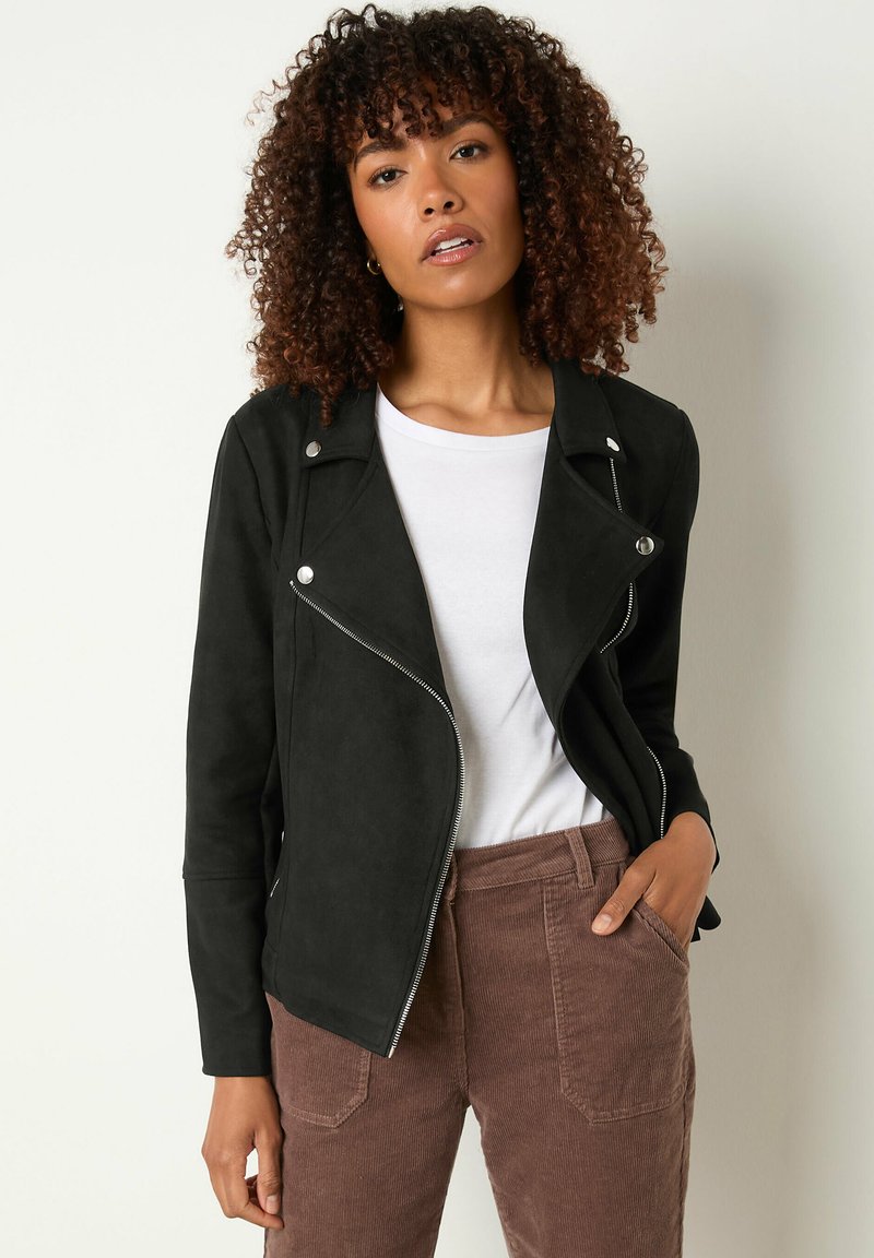 Giacca da moto in ecopelle scura con hardware argentato, zip asimmetrica e maniche lunghe; indossata sopra una camicia bianca e pantaloni in velluto a coste marroni.