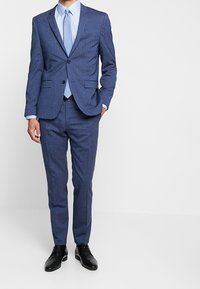Blauw pak met een jasje met twee knopen, bijpassende broek, een lichtblauw overhemd en een blauwe stropdas. Gladde stof met een subtiele textuur. Zwarte schoenen.