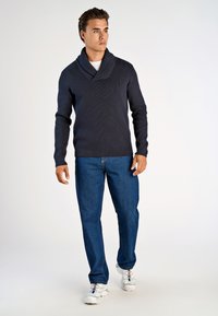 Dunkelblauer gerippter Pullover mit einem Schalkragen, kombiniert mit blauen Jeans und weißen Sneakern, die bunte Schnürsenkel haben. Einfarbiger Hintergrund.