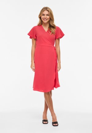 Naine seisab ja naeratab, kandes korallikarva mähkkleiti lendlevate varrukatega ja musta avatud varbaga kontsakingi valgel taustal.