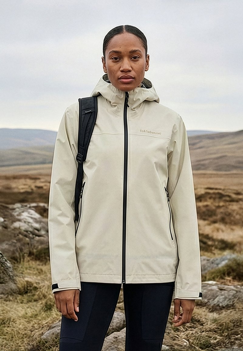 Femme portant une veste à capuche beige et un sac à dos noir, debout à l'extérieur dans un paysage rocheux et herbeux sous un ciel nuageux.