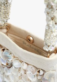 Sac en tissu beige orné de diverses coquillages. Il dispose d'une fermeture supérieure et de quincaillerie dorée décorative sur des poignées texturées. L'intérieur est doublé.