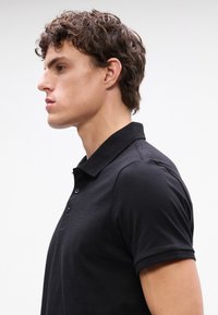 Mladý muž s kučeravými vlasmi, ktorý má oblečenú priliehavú čiernu polokošeľu, zobrazený z profilu na jednoduchom bielom pozadí.