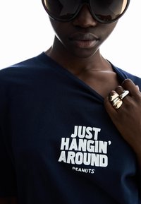 Navy cotton t-shirt featuring white text "JUST HANGIN' AROUND" in bold font. Model wears large dark sunglasses and multiple metallic rings.