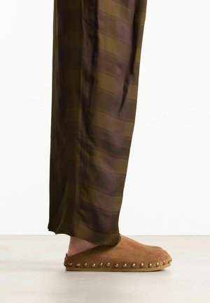 Braune Wildleder-Slipper mit runden Metallnieten entlang der Sohle, getragen zu locker sitzenden braunen karierten Hosen.