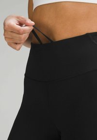 Leggings negre cu talie înaltă, cu o textură netedă. O mână ajustează o curea neagră de la talie, evidențiind un design ajustat.