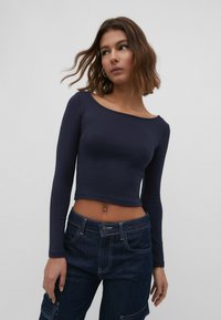 Crop top à manches longues bleu marine en tissu lisse, avec un large décolleté et une silhouette ajustée. Associé à un jean en denim taille haute.