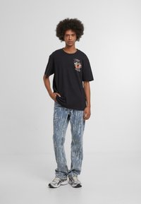 Camiseta de algodón negra con estampado gráfico en el lado izquierdo del pecho, combinada con jeans de lavado ácido azul claro con rayas blancas verticales. Zapatillas plateadas.