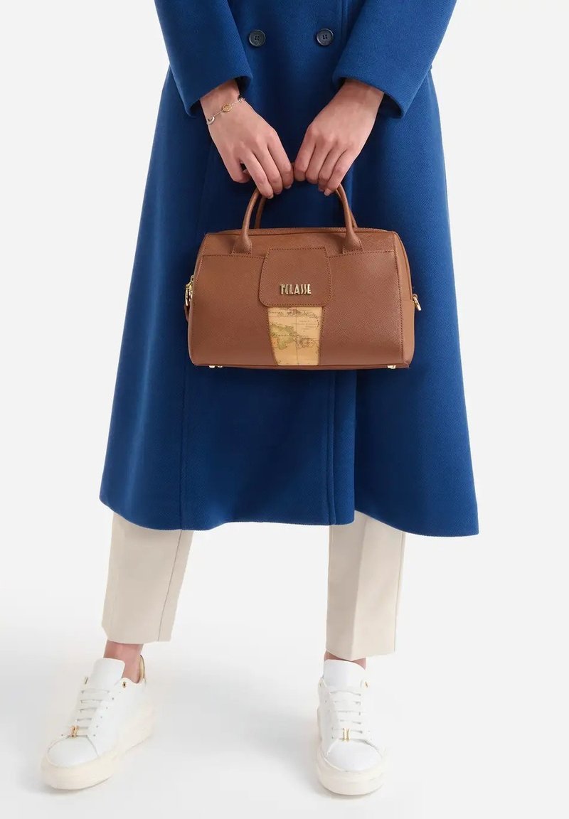 Le mani tengono una borsa in pelle marrone con dettagli a mappa, indossando un cappotto blu, pantaloni beige, sneakers bianche e un braccialetto e un anello d'oro.