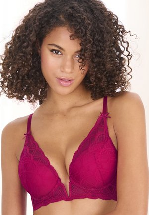 Woman with curly hair wearing a deep pink lace bra with adjustable straps and a plunging neckline, facing forward with a neutral expression.