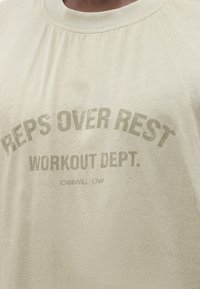 Μπλουζάκι με ξεθωριασμένο κείμενο "REPS OVER REST WORKOUT DEPT." με ανοιχτή, φθαρμένη στάμπα σε μπεζ ύφασμα, κοντινή λήψη στην περιοχή του στήθους.