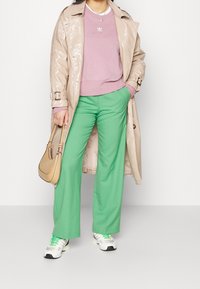 Beige blänkande trenchcoat över en rosa sweatshirt, gröna vida byxor och vita sneakers med gröna detaljer. Brun axelväska.