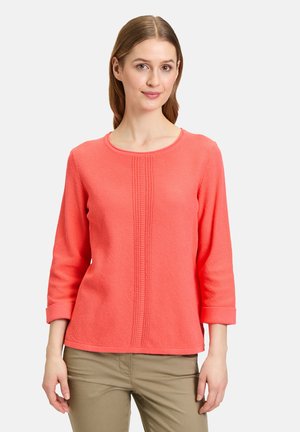 T-shirt à manches longues corail avec un col rond, en tissu texturé, et un motif central en relief. Présente des poignets roulés et une coupe décontractée.