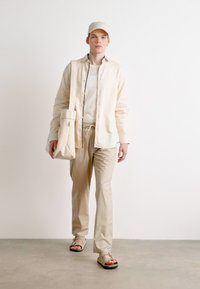 Lys beige, lett bomullskjorte over en kremfarget topp, kombinert med beige tapered bukser og flip-flops. Inkluderer en matchende caps og totebag.