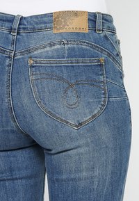 Vaqueros de denim azul con un diseño distintivo de bolsillo trasero en forma de corazón. Las etiquetas son de color marrón claro con patrones decorativos en negro. Textura suave.