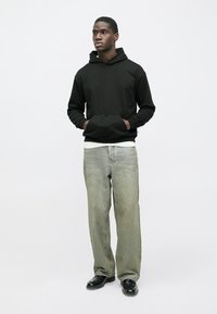 Sweatshirt preta com capuz e bolsos frontais, combinada com jeans largos verde claro. Modelo posicionado contra um fundo branco. Sapatos pretos.