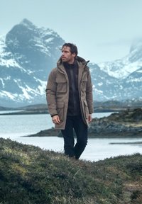 Tan parka med huva, med flera fickor, bärs över en mörk tröja. Robust terräng och snötäckta berg i bakgrunden.