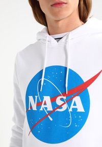 Fehér kapucnis felső húzózsinórral, kék kör alakú NASA logóval, piros swoosh szalagon és fehér felirattal, sima szövettextúrán.
