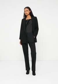 Črn oblečen blazer preko črne satenske majice in ujemajočih se hlač, ki poudarja elegantne linije, strukturirane ramena in izjemno dodelan videz.