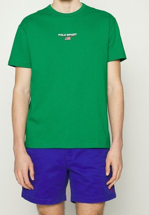 Man draagt een felgroen Polo Sport T-shirt met een klein Amerikaans vlaglogo en koningsblauwe korte broek, staand tegen een effen achtergrond.