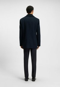 Cappotto di lana blu navy con colletto a sciallato, spalle strutturate e taglio dritto, abbinato a pantaloni scuri e scarpe, visto da dietro.