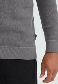 Szürke textúrájú pulóver, kötött mintával, bordás mandzsettával és hemésszel, oldalán egy kis címkével.