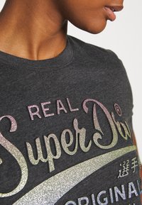 Grå bomulds t-shirt med rund hals; har glitrende tekst i pink og sølv, der står "REAL SuperDry" og "ORIGINAL."