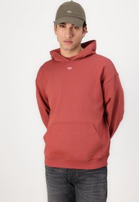 Red cotton hoodie with a front pocket and a drawstring hood, paired with dark jeans. Cap is olive green with white text.