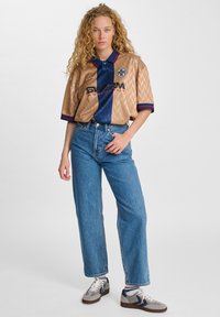 Guld- og marineblå kortærmet polo med chevron-mønster og logo, kombineret med højtaljede blå jeans og grå sneakers.