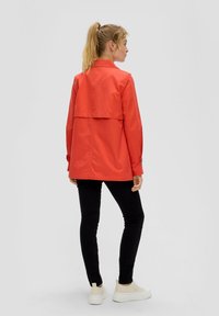 Rode jas met een achterkantflap, lange mouwen en knoopmanchetten, gecombineerd met zwarte broek en witte sneakers. Glad materiaal met een gestructureerde pasvorm.