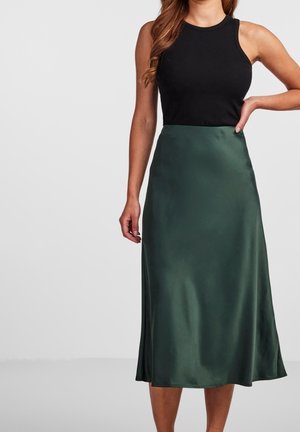 Femme portant un haut noir sans manches et une jupe midi en satin vert foncé taille haute, debout avec une main sur la hanche.