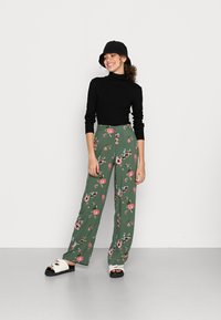 Top preto de gola alta, calças largas florais verdes com padrões em rosa e azul. Sandálias brancas com solas pretas completam o conjunto.