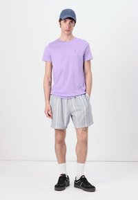 Lavendelfarbenes T-Shirt mit kurzen Ärmeln, graue gestreifte Shorts, schwarze Sneaker mit weißen Sohlen, weiße Socken und blaue Kappe. Einfacher lässiger Look.