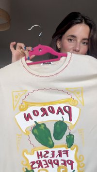 T-shirt boje kreme ima crvenu šav, okrugli izrez i grafički otisak sa zelenim paprikama i rečju "PADORON" u podebljanoj crvenoj boji.