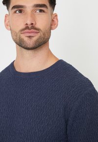 Granatowy sweter z teksturowanym wzorem w diamenty, okrągłym dekoltem i miękkim materiałem. Zbliżenie na górną część ciała, prezentujące szczegóły wzoru.