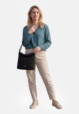 Femme blonde portant un chemisier bleu sarcelle et un pantalon beige, tenant un sac à main noir avec une bandoulière, debout devant un fond uni.