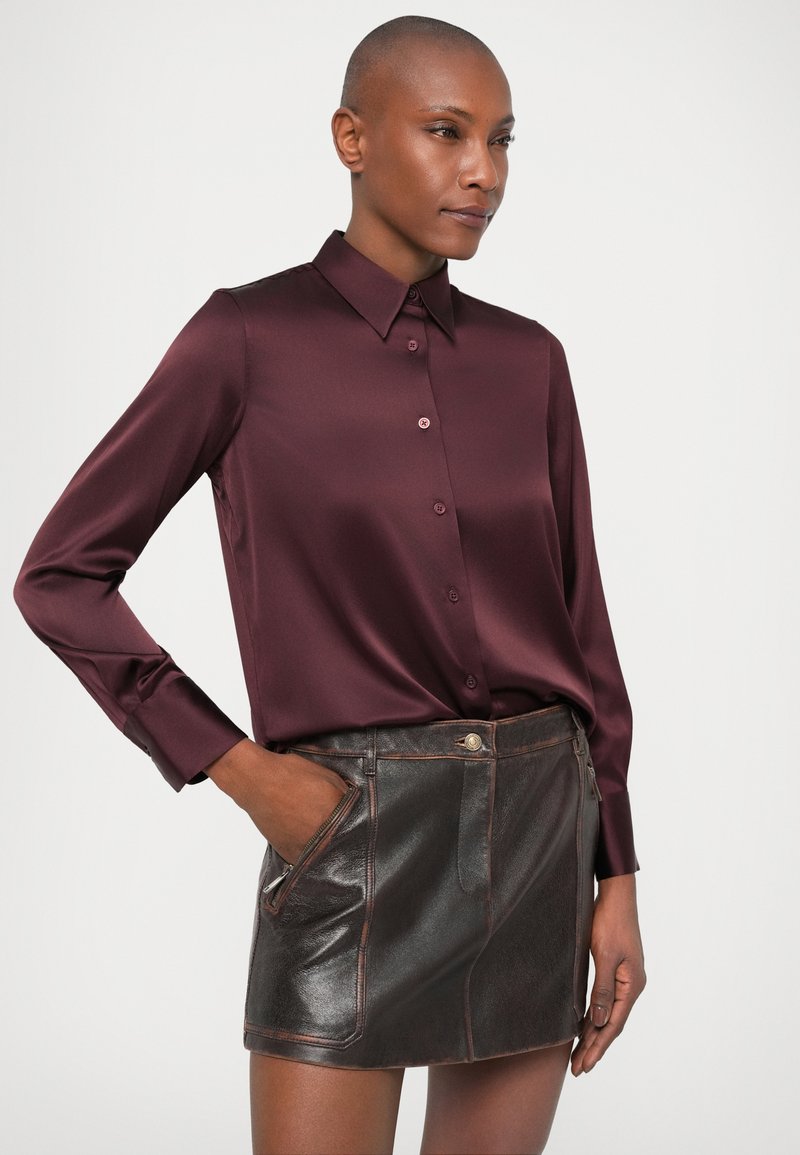 Frau in einem bordeauxroten Satin-Hemd mit langen Ärmeln, das in einen dunkelbraunen Leder-Minirock mit Reißverschlusstaschen gesteckt ist.