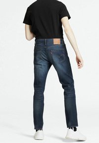 Jeans de mezclilla azul oscuro con corte ajustado, que cuentan con un parche de cuero marrón en la cintura y zapatillas blancas, vistos desde la parte trasera.
