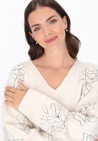 Kremowy sweter z dekoltem w serek z czarną haftowaną florystyką i cekinowymi akcentami na rękawach. Miękka struktura dzianiny; ściągacze na rękawach i u dołu.