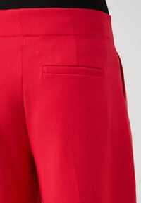 Red tailored trousers with a smooth texture, featuring a back pocket and subtle stitched details along the seams.