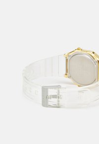 Bracelet de montre en plastique transparent avec boîtier en métal doré, forme octogonale, dos argenté et boucle grise avec "Casio" gravé.