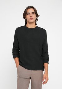 Zwarte gebreide sweater met een ronde halslijn en opgerolde mouwen, met een gestructureerd oppervlak. Gecombineerd met lichtbruine broeken.