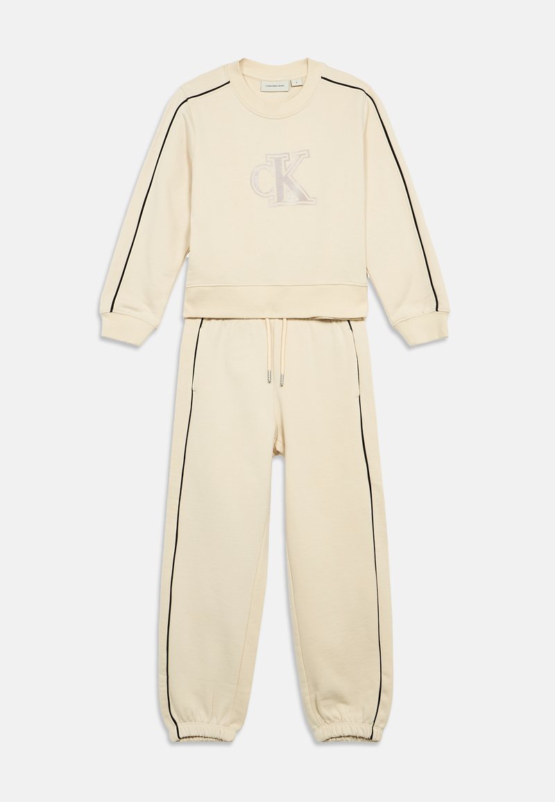 Creme sweatshirt en joggers set, met zwarte zijstrepen en een tonale geborduurde logo op de borst. Zacht materiaal met elastische boorden.