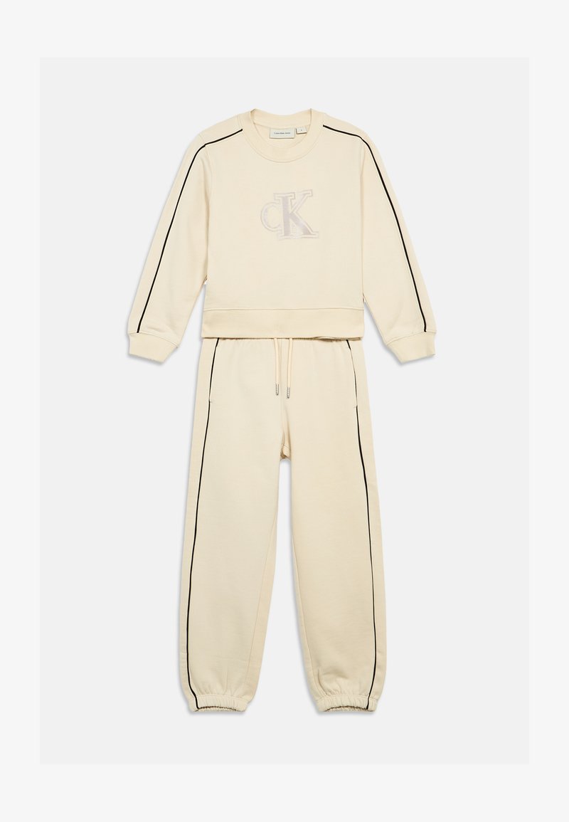 Krēma krāsas sporta krekls un bikses komplekts, ar melnām sānu svītrām un tonālām izšūtām logo uz krūšu daļas. Mīksta auduma materiāls ar elastīgiem aprocēm.