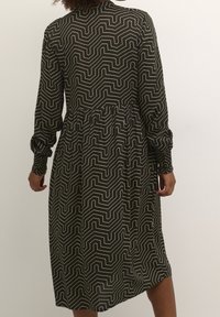 Robe à manches longues en tissu vert foncé avec un motif géométrique en zigzag blanc, dotée d'une taille cintrée et d'une jupe fluide.