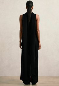 Černé bezrukávové šaty s volným siluetou, které mají otevřená záda a boční rozparky, vyrobené z lehkého materiálu.